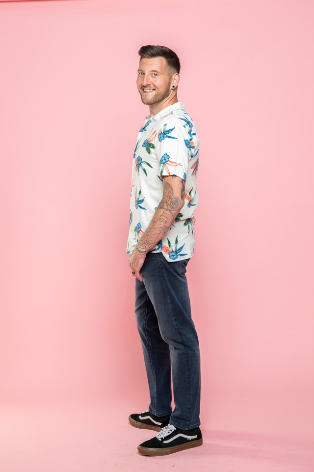 Man in floral shirt and jeans smiling against pink background.