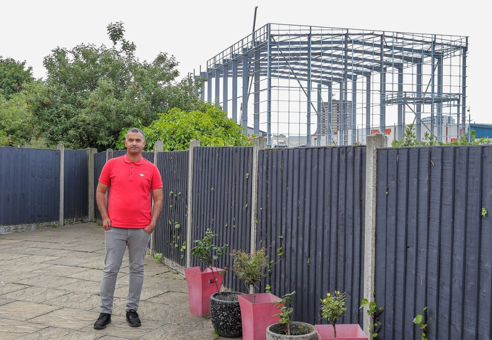Man in red shirt standing in his garden next to a fence, with a large metal warehouse frame under construction visible in the background.