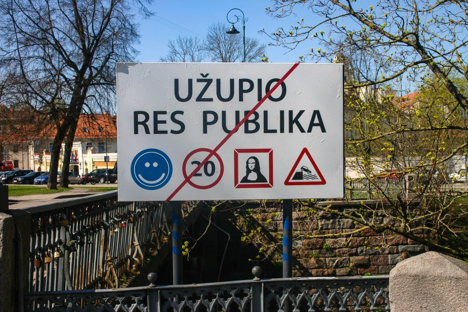 Sign indicating Užupio Res Publika with a smiley face, a crossed-out 20 speed limit, Mona Lisa, and a downhill traffic sign.