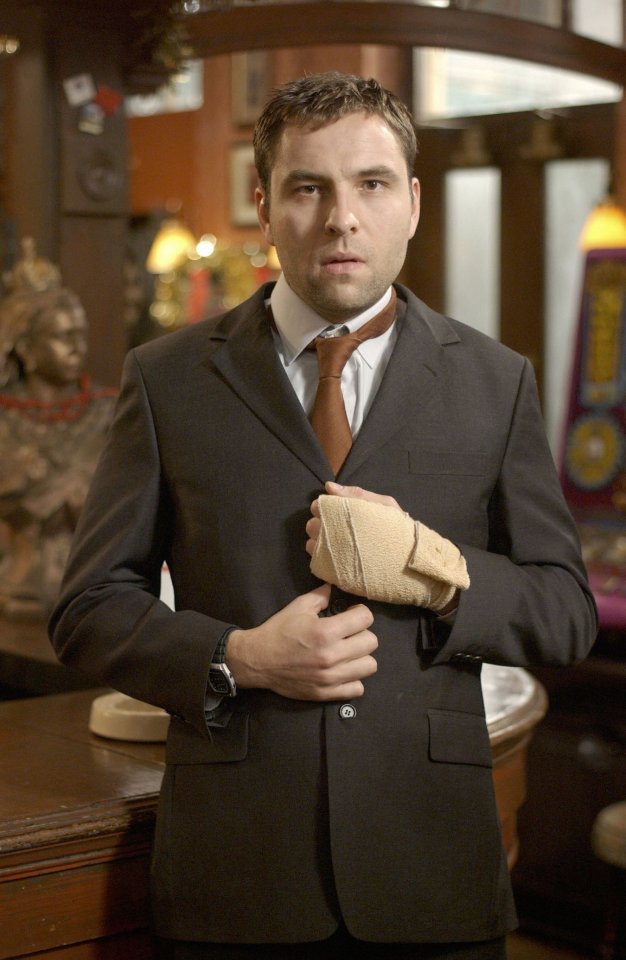 David Walliams as Ray in EastEnders, wearing a suit and a bandage on his hand.