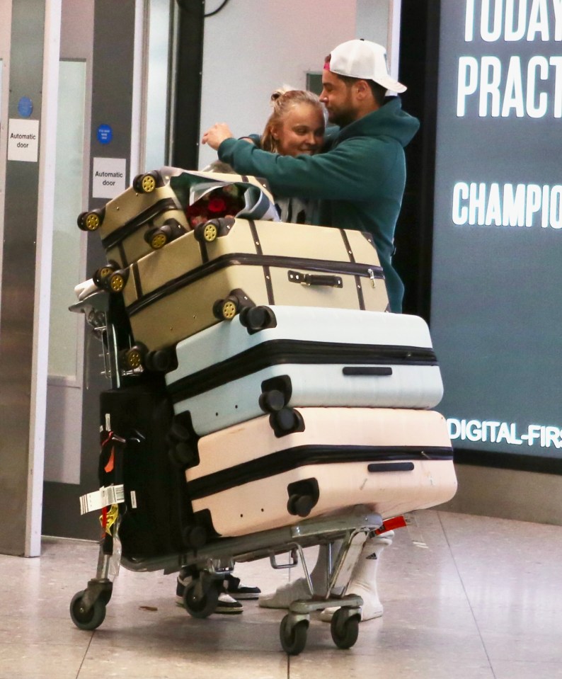 Jojo Siwa and Chris Hughes embracing at an airport, luggage cart in front of them.