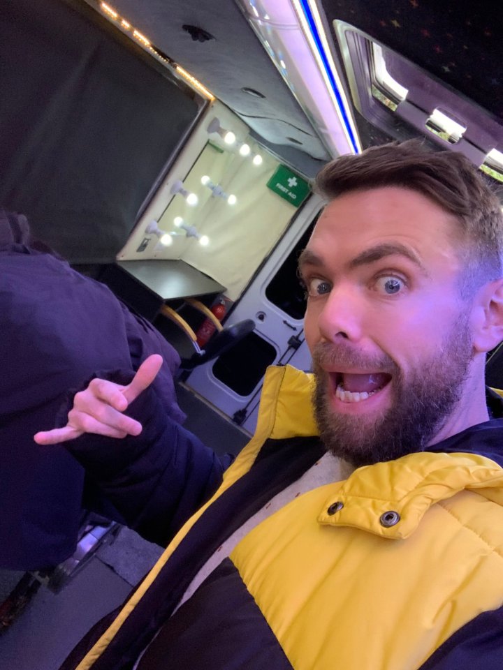 Man in a yellow jacket making a hand gesture inside a van with a makeup station.