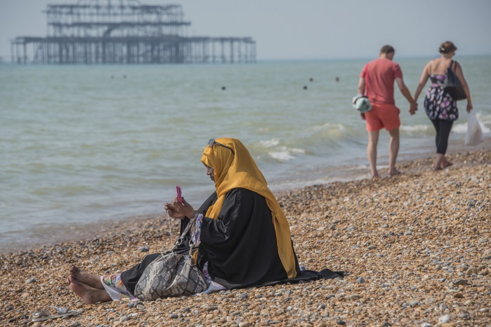 A mother, in a hijab, watches over her family who are also modestly dressed, as they play in the sea on Brighton Beach