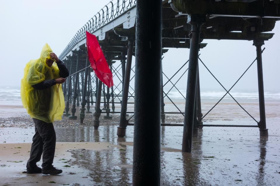 Man walking under Saltburn's Victorian pier struggles with umbrella on a wet and windy September morning. Saltburn, England, UK