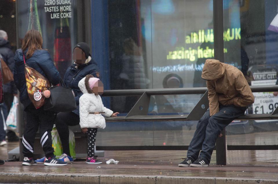  A man under the influence of the drug Spice is watched by confused girl queuing for the bus with her family