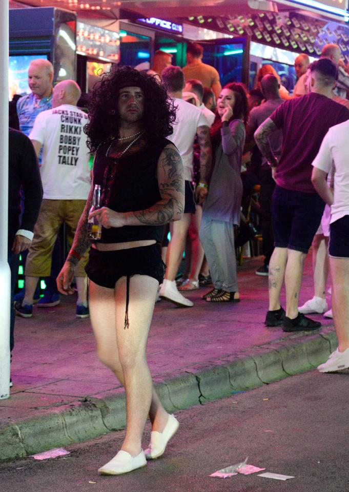  A tattooed man wearing a wig and small shorts clutches a beer as he walks down the main strip of clubs and bars