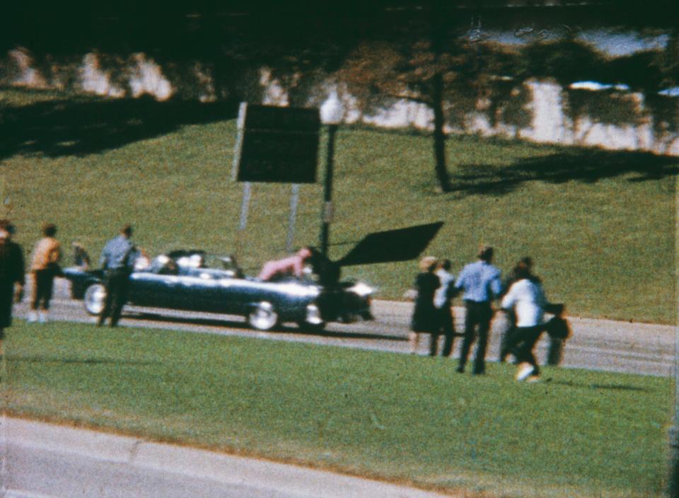  Bystanders look on as Jacqueline Kennedy reaches over to help husband John F. Kennedy who lies on the rear of a car after being struck by an assassin's bullet
