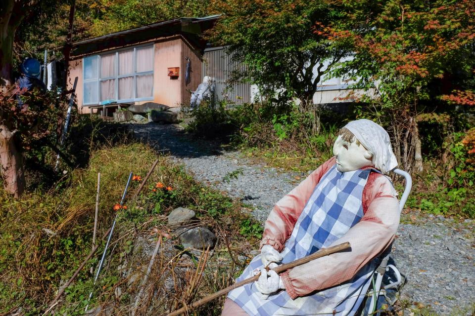  An elderly woman doll sits in a driveway wearing an apron and shawl with a walking stick in her hands
