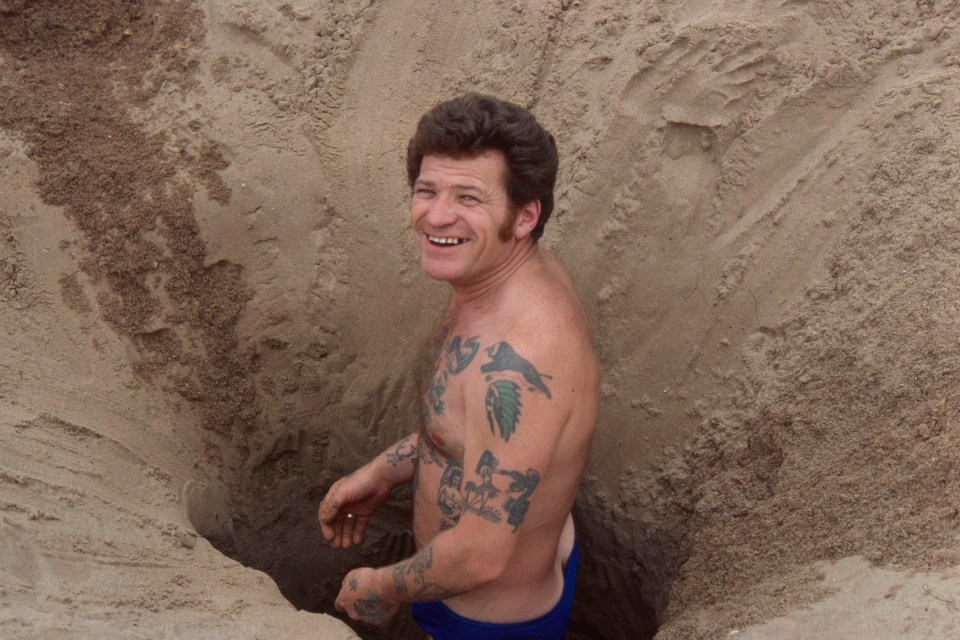 A man grins from an enormous hole he's dug in the sand on a British beach