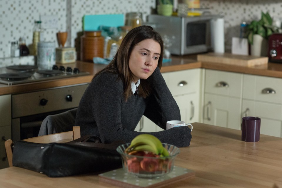 A young woman sits at a kitchen table, looking thoughtful.