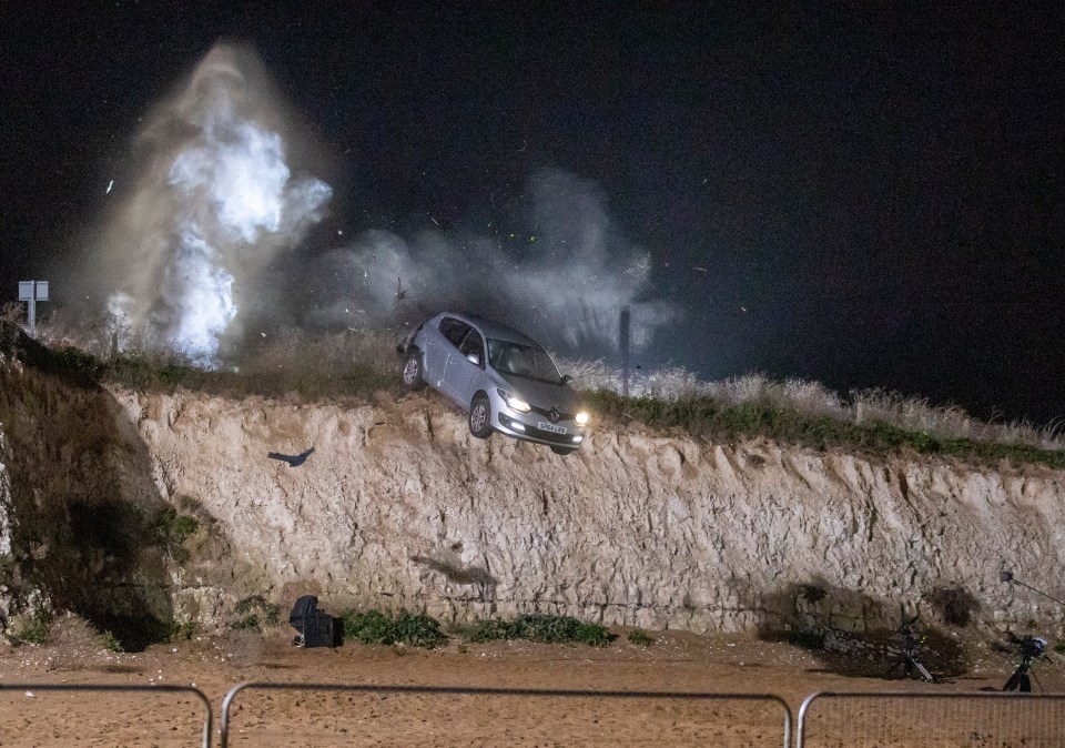 The car hurtles off the cliff before Mick appears to follow it