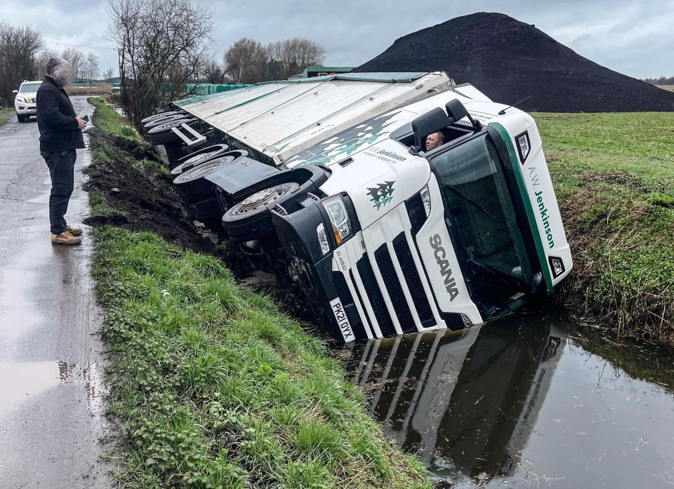 A lorry driver peered helplessly out of his trick as it lies on its side in a ditch
