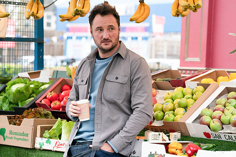 Martin Fowler from EastEnders at a fruit and vegetable stand, holding a coffee cup.
