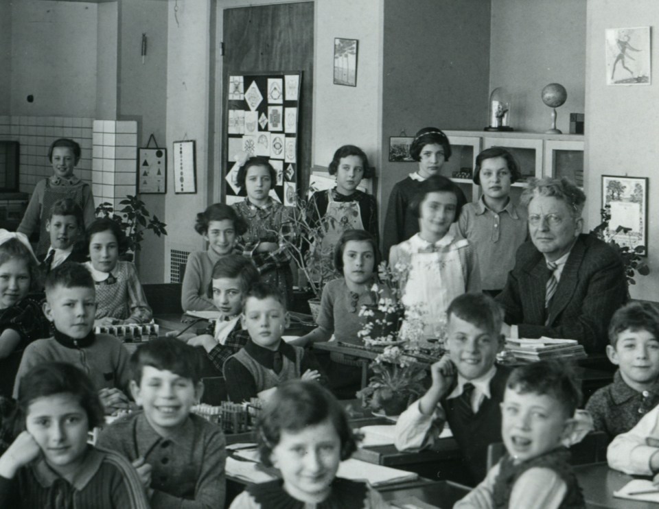 Hannah and Anne in their classroom at the 6th Montessori school in Amsterdam in 1938. Hannah is on the far left, seated towards the back, and Anne stands in a white pinafore to the left of their teacher