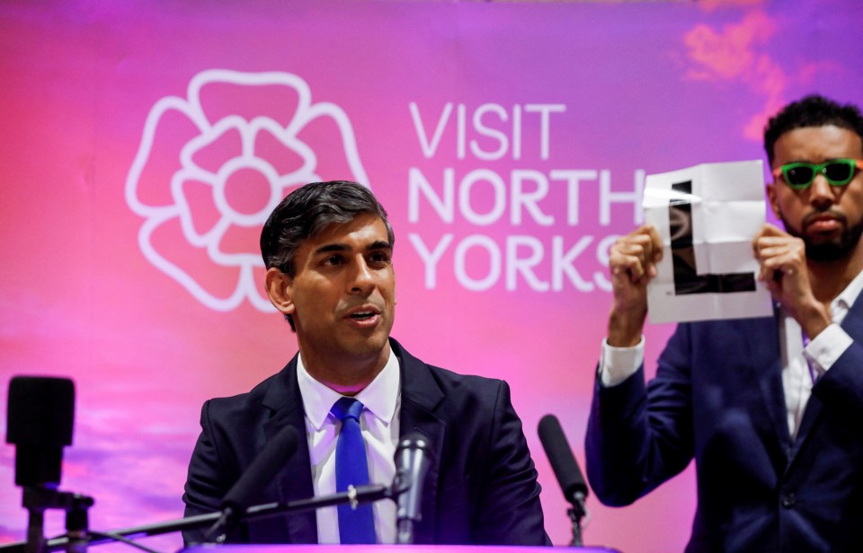Rishi Sunak speaking after winning his seat in the UK general election, with a man holding up an "L" behind him.