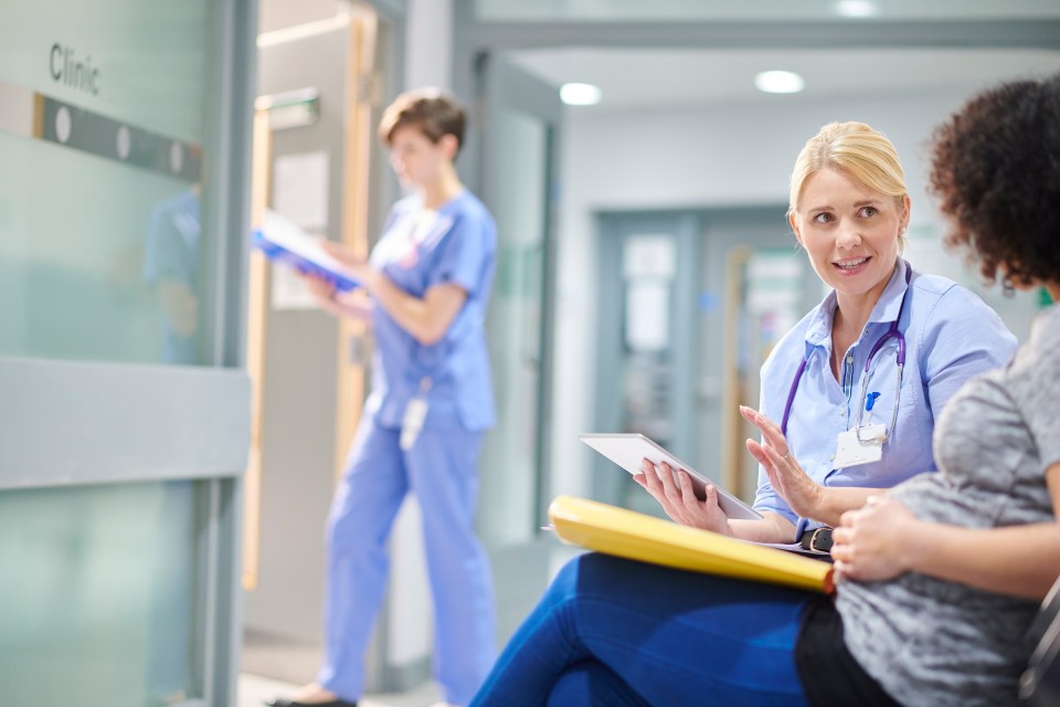 a pregnant woman waiting in hospital , sitting in the waiting room and chatting to a doctor with a young nurse in the background.