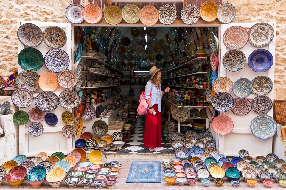 Young blonde woman in a long red skirt, hat and with pink backpack is standing in a small shop and looking at Moroccan colorful plates.; Shutterstock ID 1058262620; purchase_order: -; job: -; client: -; other: -