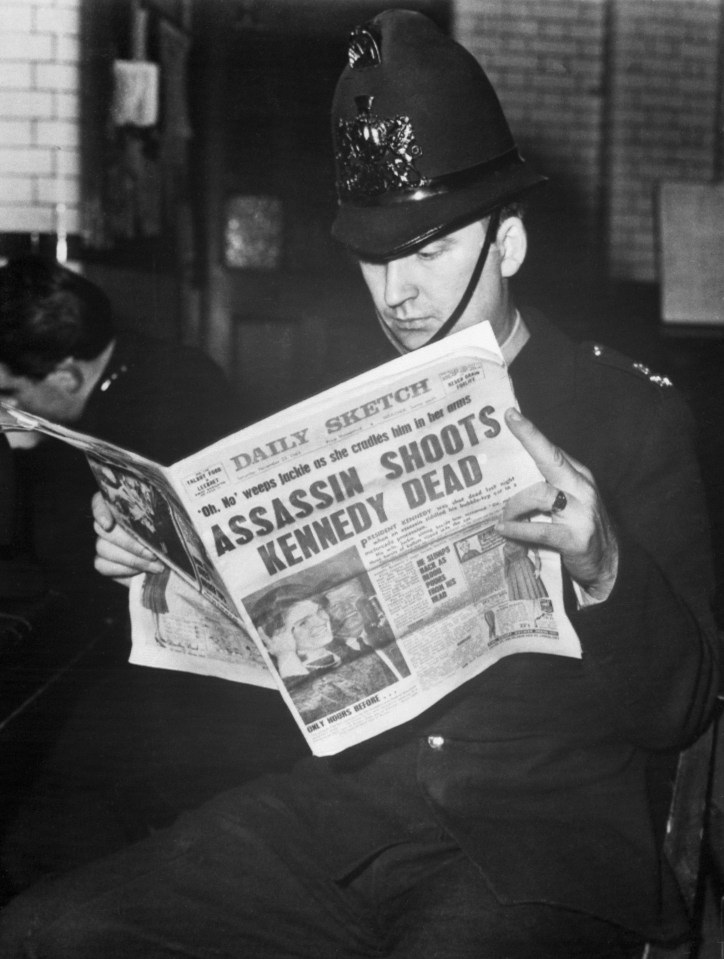 A police officer in London (a "bobby") reads the November 22, 196, issue of the Daily Sketch. The headlines proclaim: "Assassin Shoots Kennedy Dead".