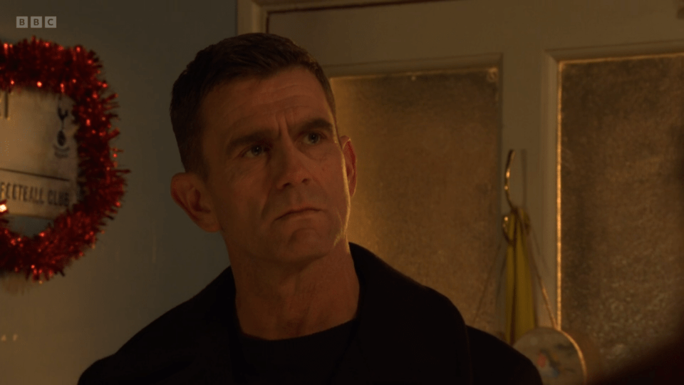 A man looking pensive, standing in a hallway with a Christmas wreath in the background.
