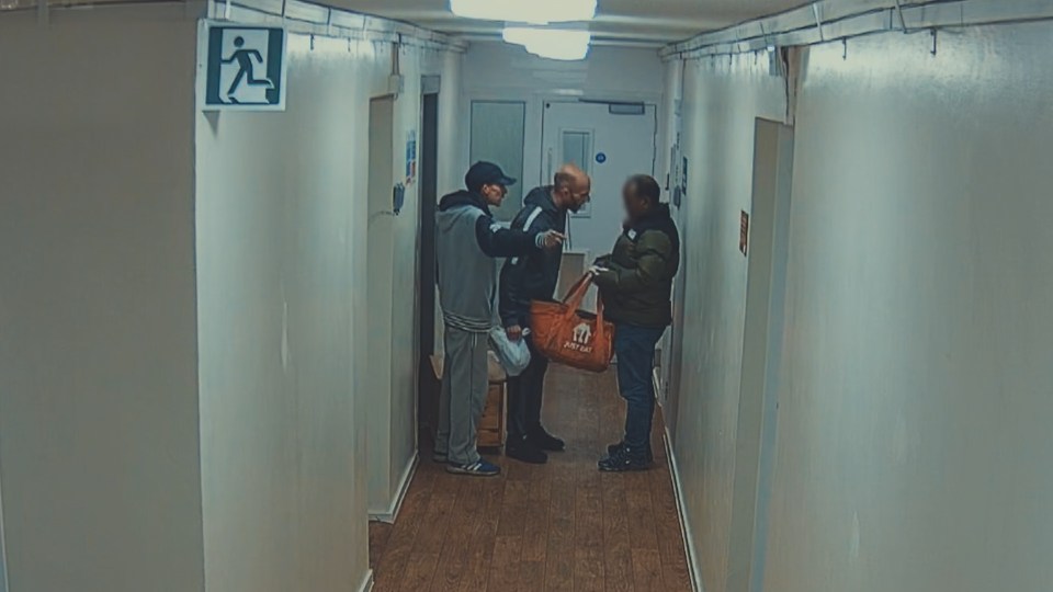Three men arguing in a hallway, one holding a Just Eat delivery bag.