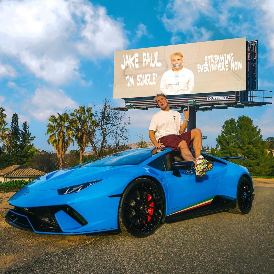 a man sits in a blue sports car in front of a billboard for jake paul
