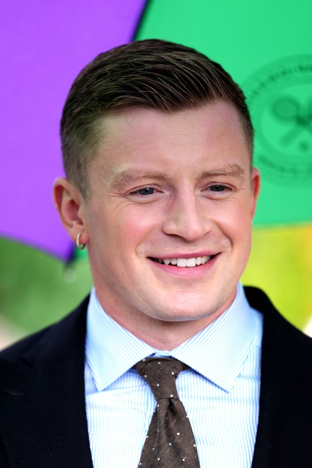 a man in a suit and tie smiles in front of a purple umbrella