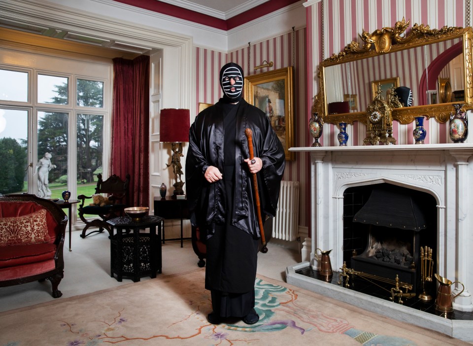 a man in a black robe and mask holds a cane in front of a fireplace