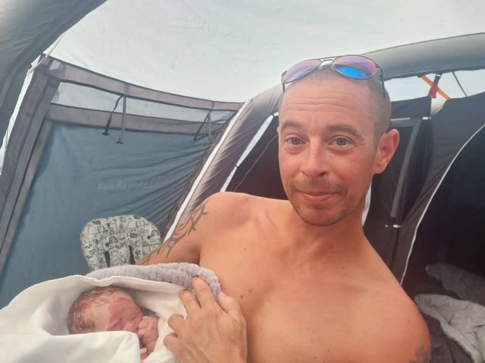 a shirtless man holds a baby in front of a tent