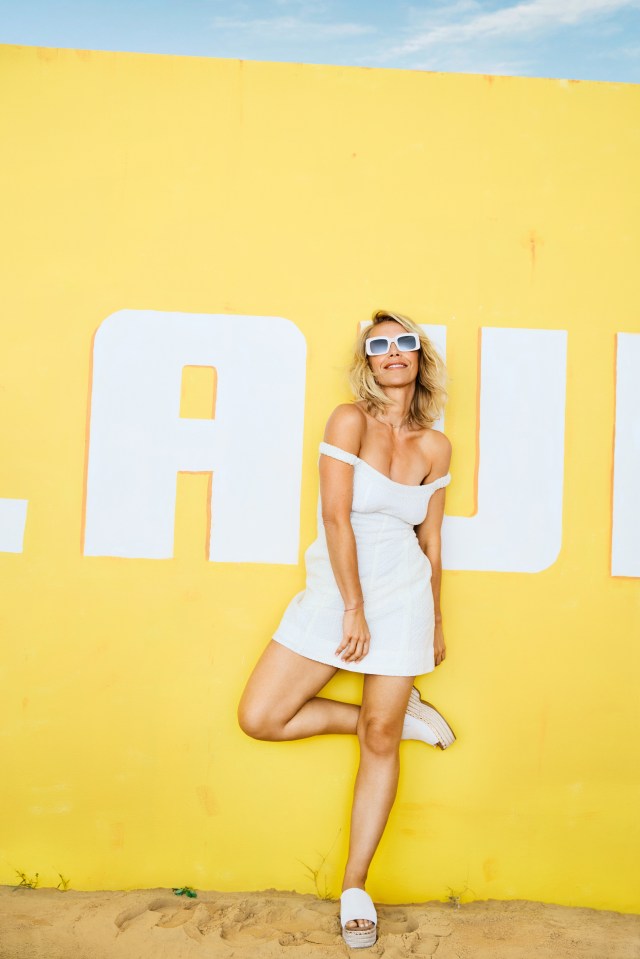 a woman in a white dress leans against a yellow wall that says au