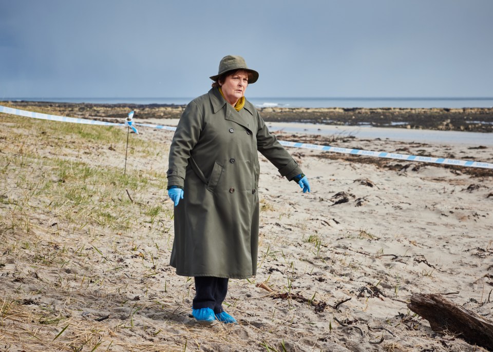 a woman in a trench coat and hat stands on a sandy beach