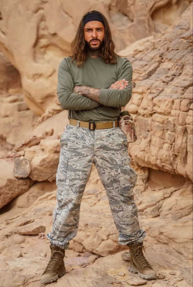 a man with long hair and a beard is standing in the desert with his arms crossed