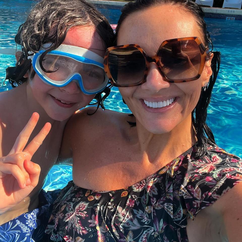 a woman wearing sunglasses and goggles stands next to a boy wearing goggles and giving a peace sign