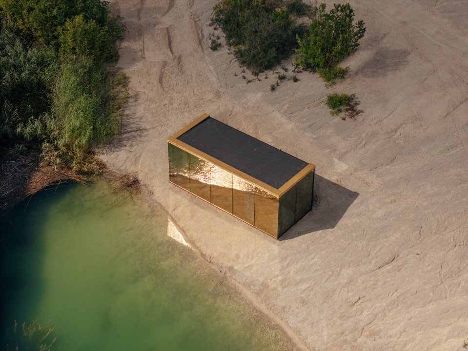 a small building with a black roof sits in the sand near a body of water