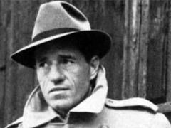 a black and white photo of a man wearing a hat and trench coat .