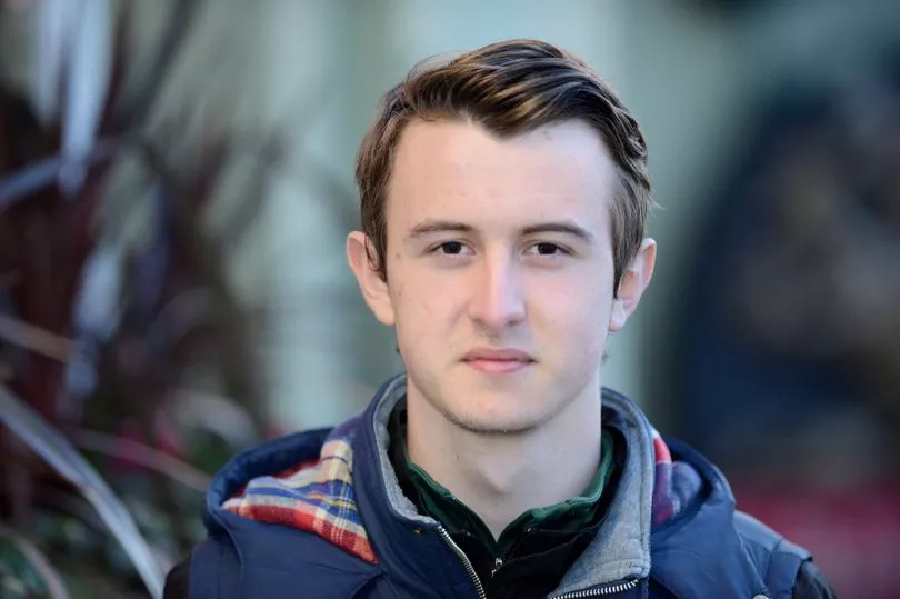 a young man wearing a plaid shirt and a blue vest looks at the camera