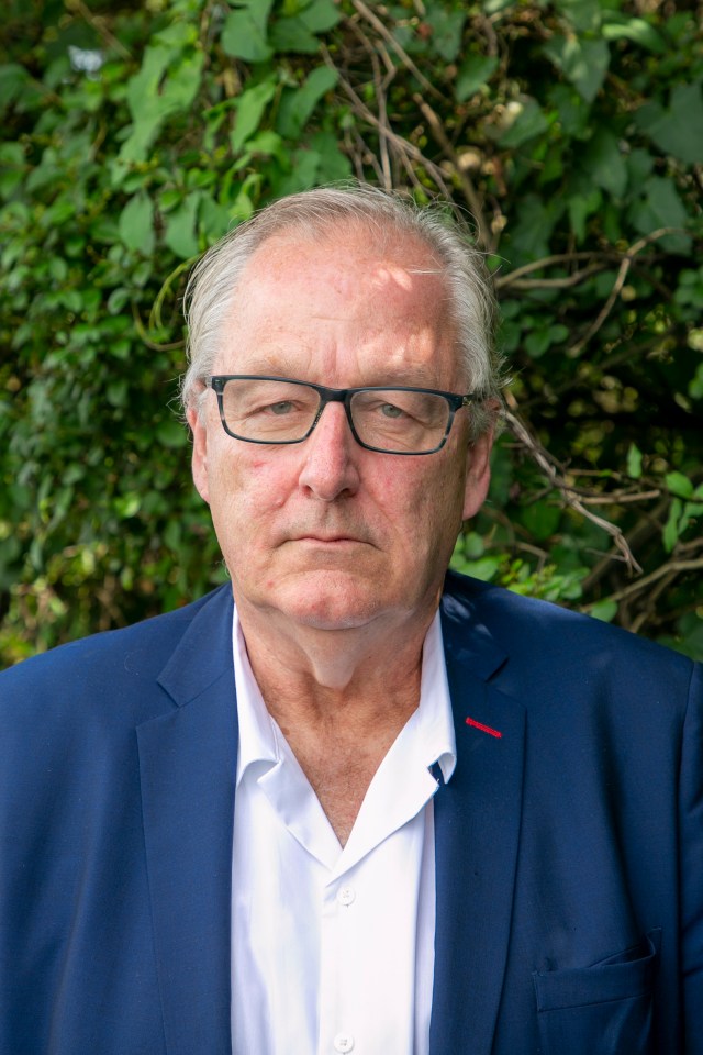 a man wearing glasses and a blue suit stands in front of a bush