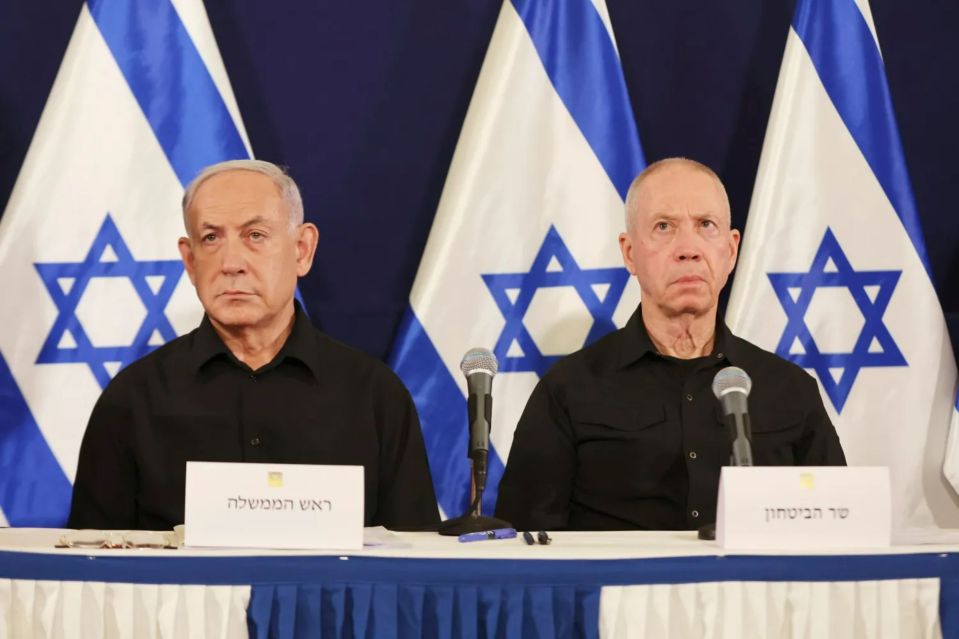 two men sit at a table with microphones in front of flags with a star on them