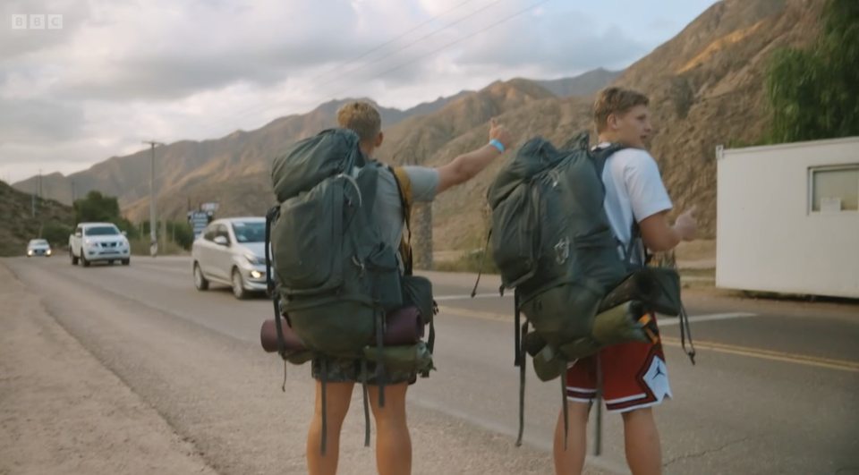 two men with backpacks hitchhiking on the side of the road