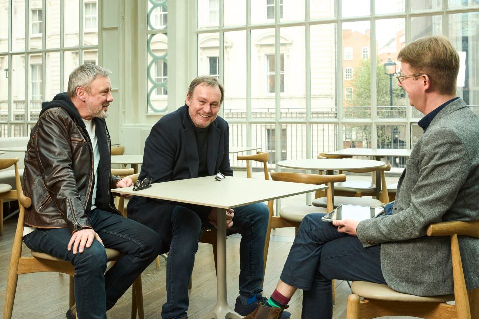 three men are sitting at a table in front of a large window