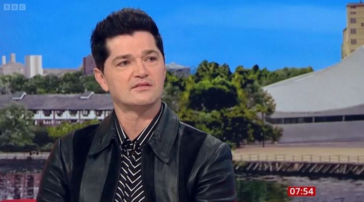 a man wearing a leather jacket and a striped shirt is talking on a television show .