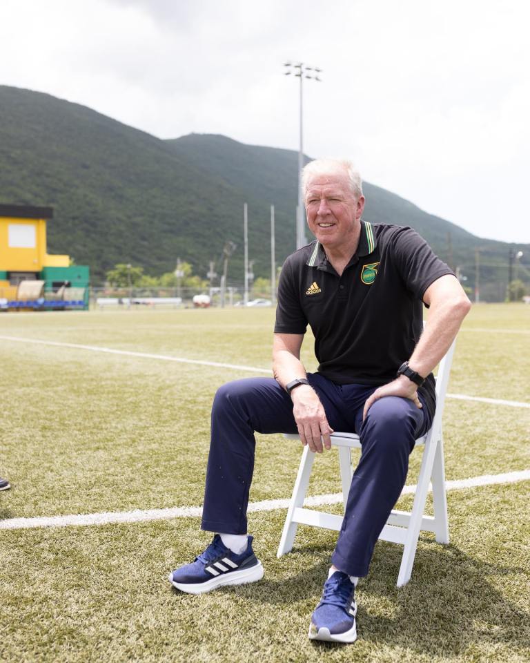 a man wearing an adidas shirt sits on a chair on a soccer field