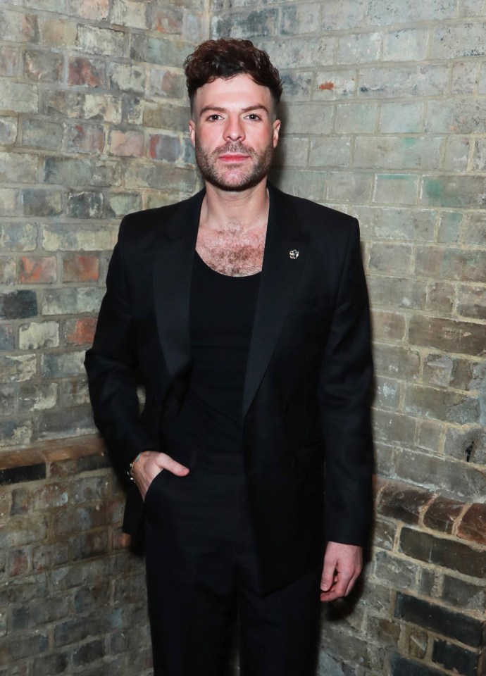 a man in a black suit stands in front of a brick wall