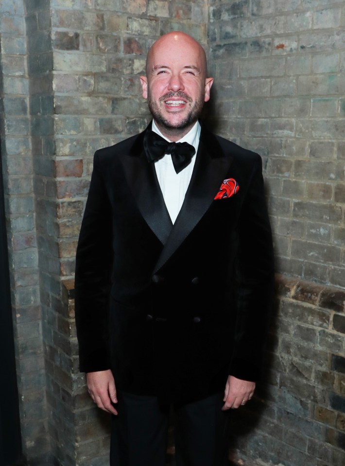 a man in a tuxedo and bow tie smiles in front of a brick wall
