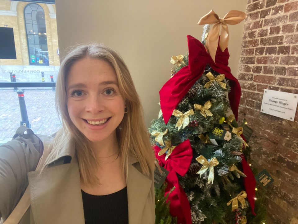 a woman stands in front of a george singer christmas tree