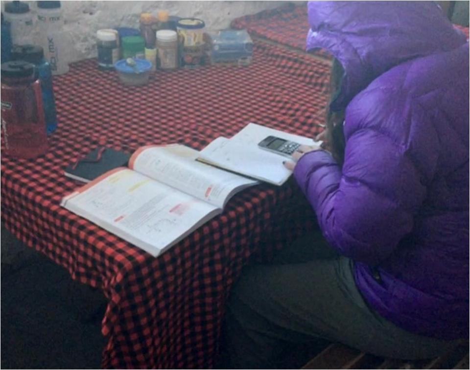 a person in a purple jacket sits at a table reading a book and using a calculator