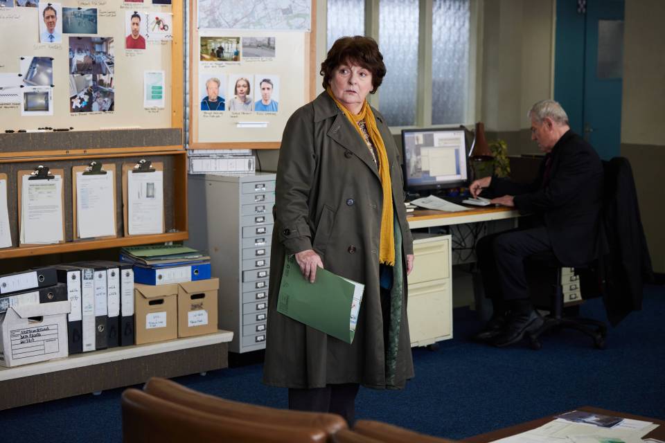 a woman in a trench coat is standing in front of a bulletin board with pictures of people on it