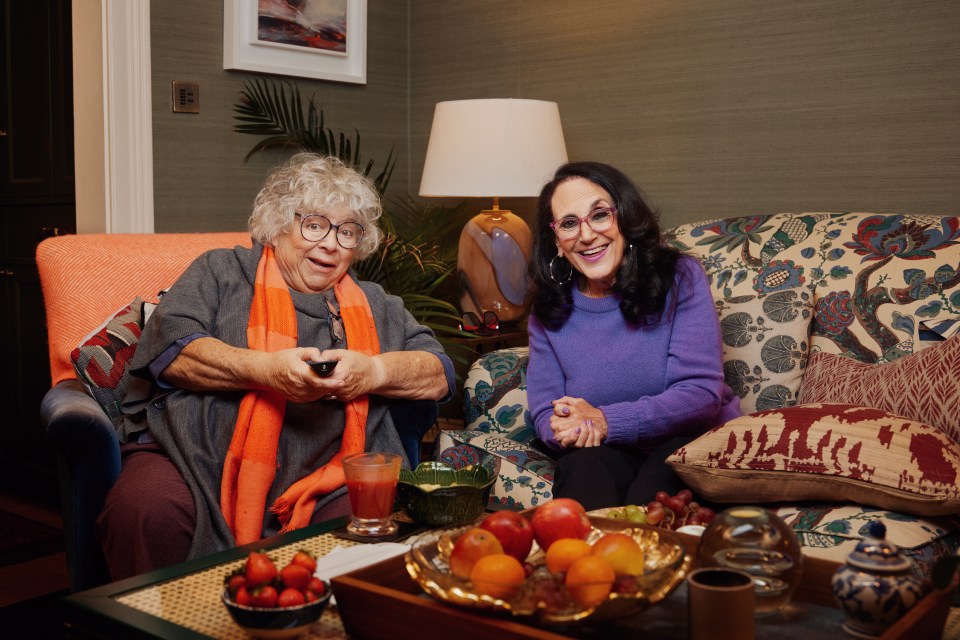 two women sit on a couch with a bowl of fruit on the table