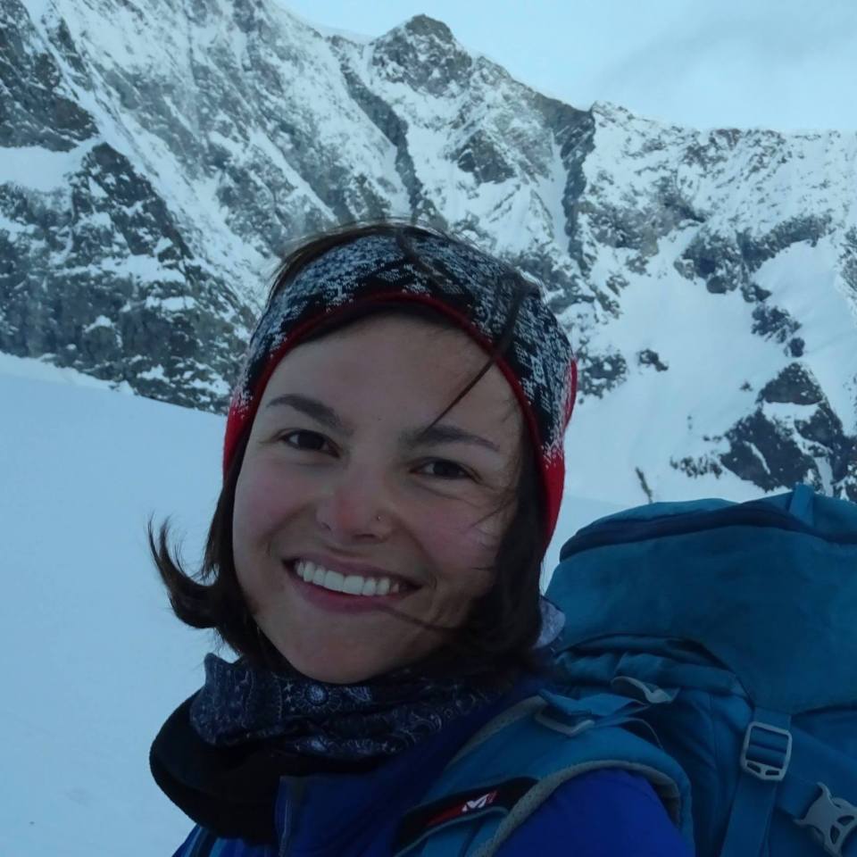 a woman wearing a blue millet backpack smiles in front of a snowy mountain