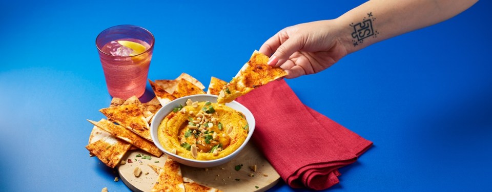 a person with a tattoo on their arm dipping a tortilla chip into a bowl of hummus