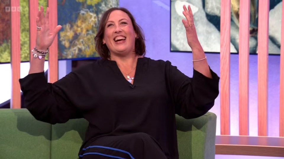 a woman sits on a green couch with her arms in the air and a bbc logo behind her